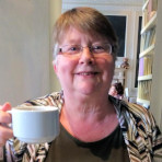 Colour photo of head and shoulders of Elaine, smiling, indoors, and raising a cuppa to the camera