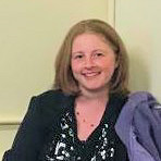 Colour photo of head and shoulders of Jessica, smiling, wearing choir uniform of a black long sleeved shirt, trousers, and on this occasion, a black scarf patterned with silver musical notes