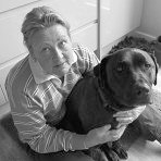 Greyscale photo of Jane seated on the floor indoors, holding her dog; looking upwards towards the camera