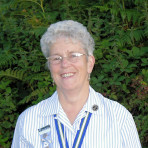 Colour photo of head and shoulders of A A, smiling, wearing a blue and white striped blouse; with green foliage in the background