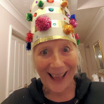 Colour headshot of Celia, smiling; wearing a tall-hat creation, with gold foil bands, studded all over with coloured tinsel pom-poms and gem-stones