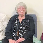 Colour photo of Pam, smiling, sitting indoors, wearing choir uniform of a black long sleeved shirt, trousers, and on this occasion, a black scarf patterned with silver musical notes