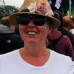 Colour photo of head and shoulders of Rebecca, smiling, at a music festival, wearing a white T-shirt, sunglasses, and a straw sun-hat decorated with flowers